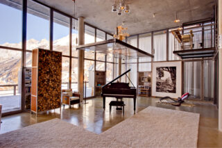 Firewood storage and grand piano beneath raised suspended table in the living space at the Heinz Julen Loft in Zermatt